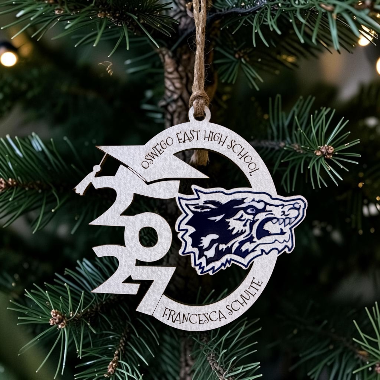 A personalized metal Christmas ornament hanging from a tree branch. The ornament is silver-colored with a custom design featuring the text 'OSWEGO EAST HIGH SCHOOL', '2027', Senior Name', and a navy blue and white wolf mascot logo.
