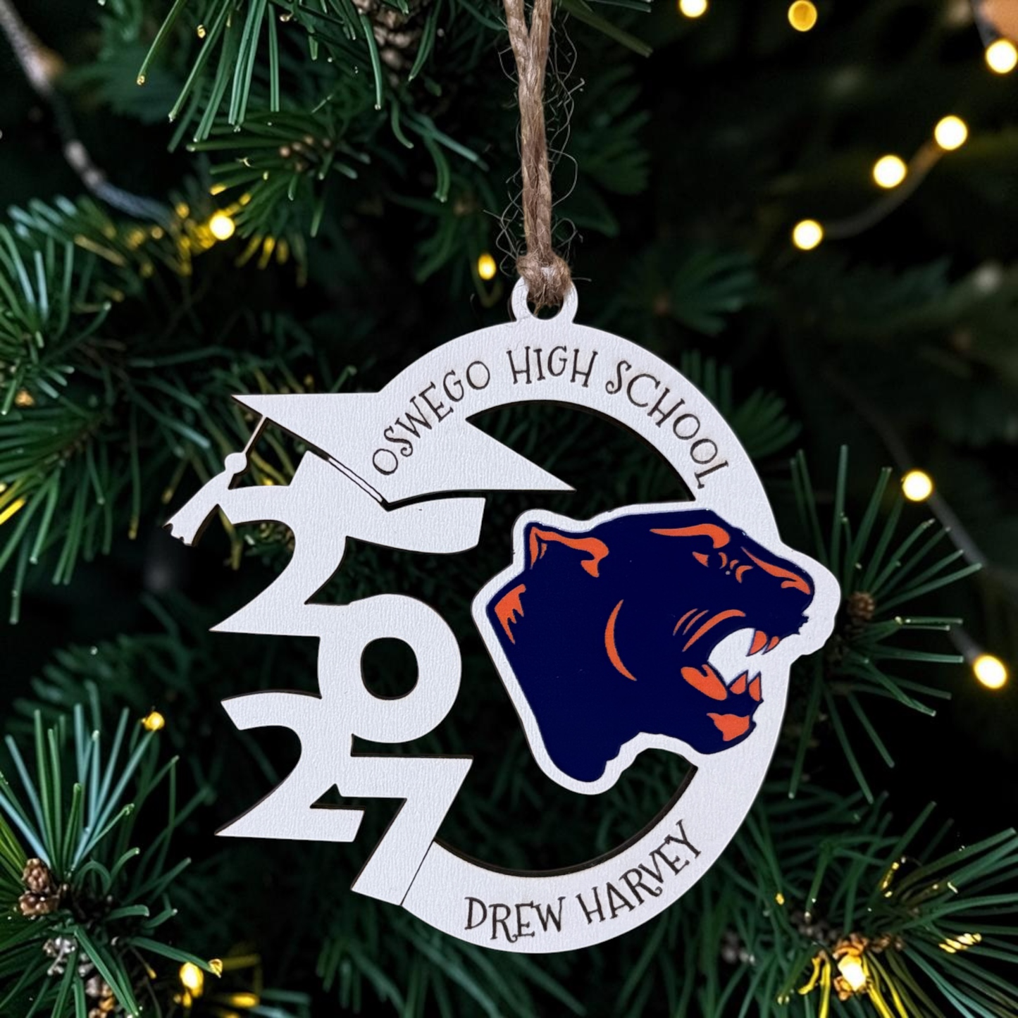 A personalized school Christmas ornament hanging on a Christmas tree. The circular white ornament features a navy blue and orange wolf mascot logo, the text 'OSWEGO HIGH SCHOOL' and 'DREW HARVEY', and the year '2027' stylized with a graduation cap and lightning bolt.
