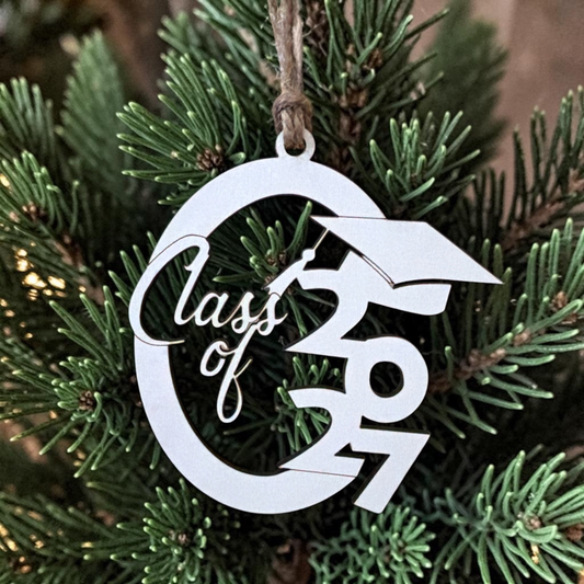 A white hardboard Christmas ornament shaped like a graduation cap and the words 'Class of 2027', hanging from a twine loop against green pine tree branches.