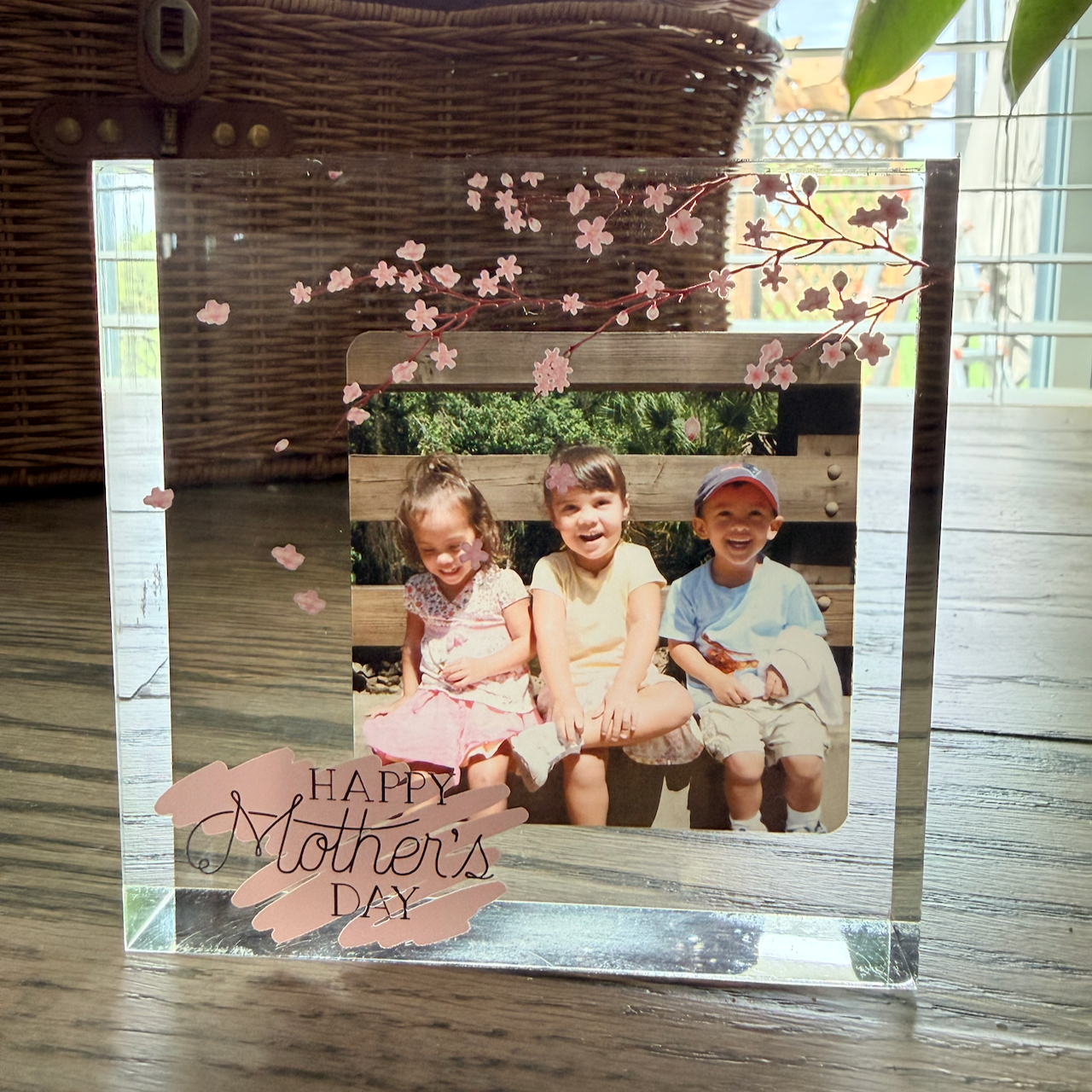 Custom acrylic photo block with cherry blossom design and Happy Mother's Day text, displaying a photo of three children sitting on a bench. Shown on a wood surface with a wicker basket in the background.