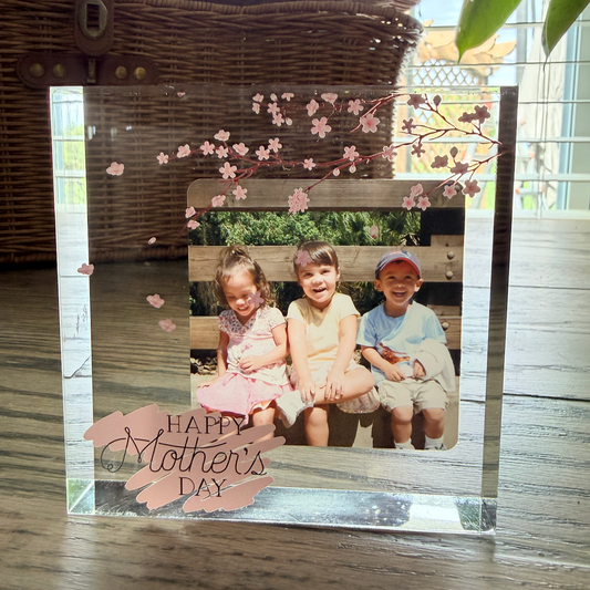 Custom acrylic photo block with cherry blossom design and Happy Mother's Day text, displaying a photo of three children sitting on a bench. Shown on a wood surface with a wicker basket in the background.