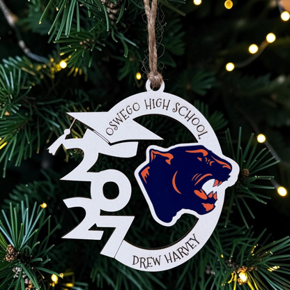 A personalized school Christmas ornament hanging on a Christmas tree. The circular white ornament features a navy blue and orange wolf mascot logo, the text 'OSWEGO HIGH SCHOOL' , and the year '2027' stylized with a graduation cap and lightning bolt.