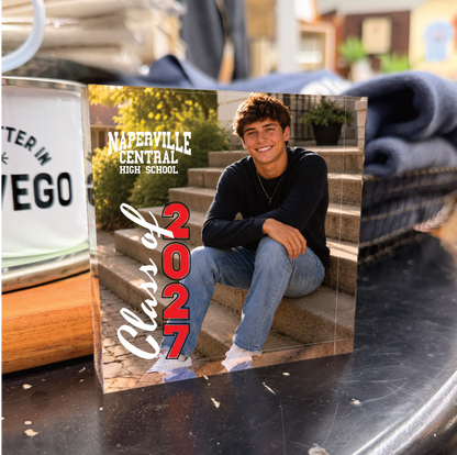 A custom square glass or acrylic photo block featuring a printed photo of a young man sitting on steps, with the text 'Naperville Central High School' and 'Class of 2027'. The block is displayed on a dark reflective surface next to a white metal mug.