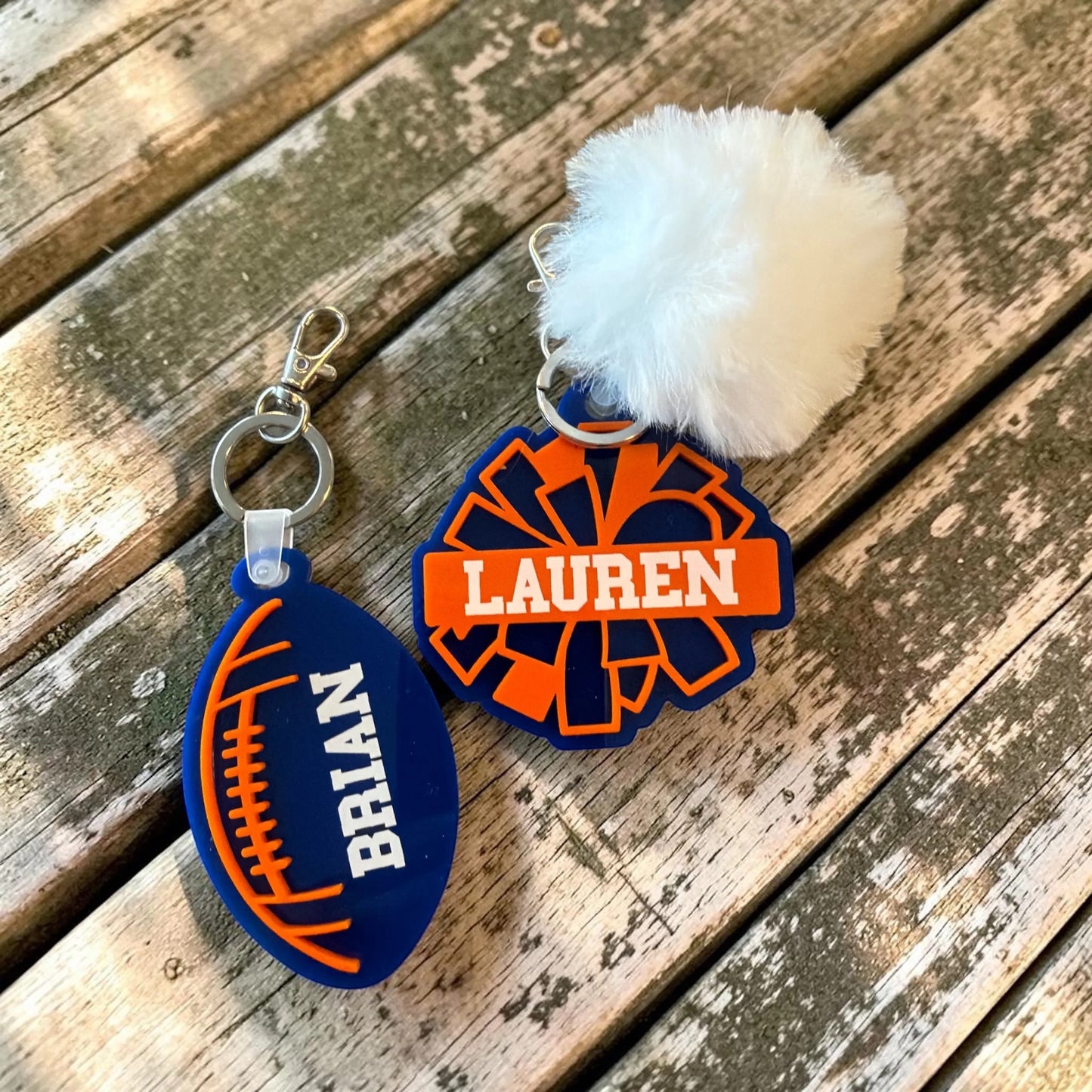LZeeDoodle Designs personalized acrylic football and cheer pom-pom bag tags in blue and orange on a wooden surface.